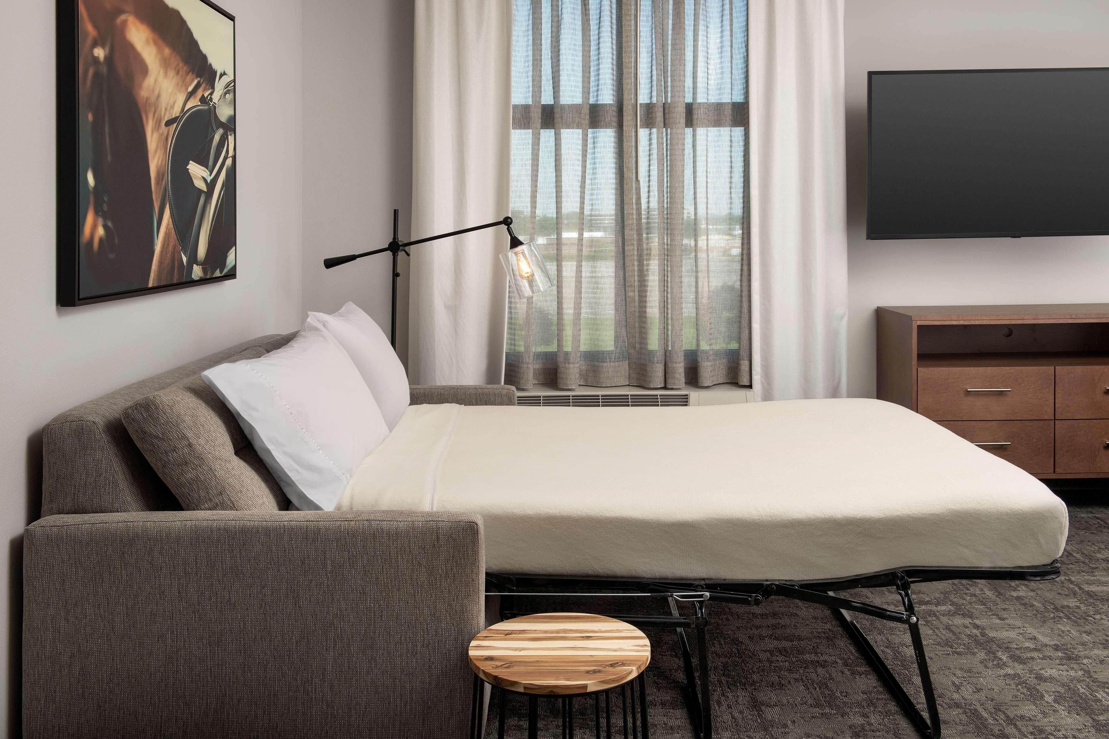 Una habitación de hotel moderna con cama, una silla, un taburete de madera, televisor y ventana con cortinas.