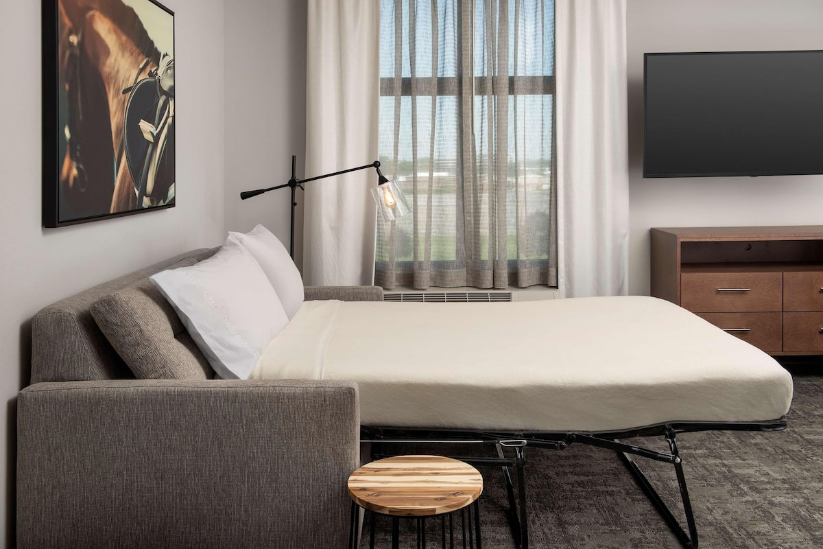 A modern hotel room with a bed, a chair, a small wooden stool, a TV, and a window with curtains.