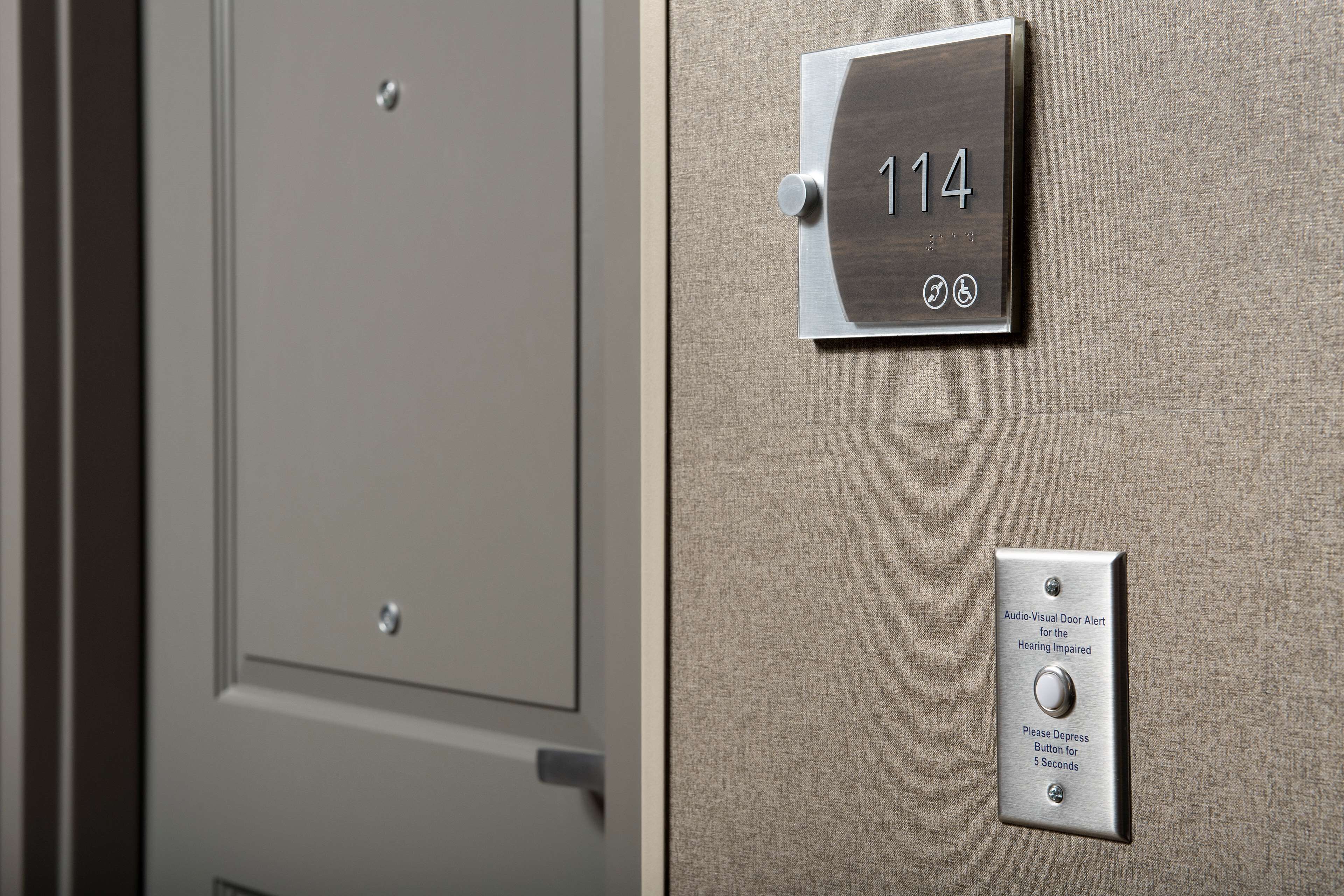 Uma porta com placa de identificação com o número "114" e um botão de alerta audiovisual para deficientes auditivos.