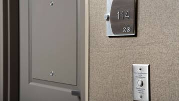 Uma porta com placa de identificação com o número "114" e um botão de alerta audiovisual para deficientes auditivos.
