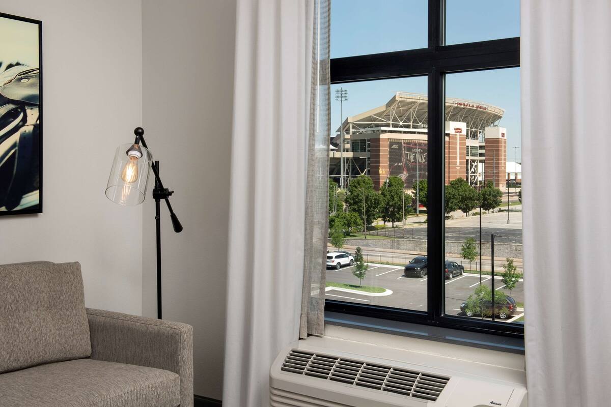 A hotel room with a large window, a sofa, a floor lamp, and a wall-mounted air conditioner.