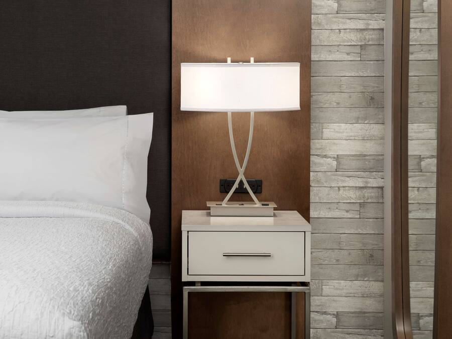 Una cama bien tendida con ropa blanca, una mesita de noche con una lámpara y una pared de paneles de madera.