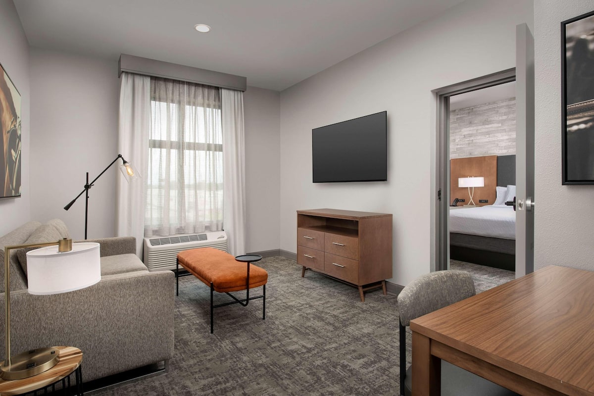 A modern hotel room with a grey sofa, a wooden table, a desk with a lamp, a wardrobe, a bed with a headboard, and a large flat-screen TV.
