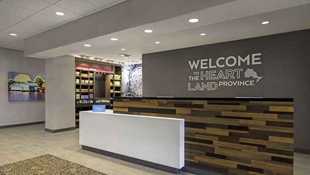 A reception desk with a "Welcome to the Heartland Province" sign, a hotel directory, and a wall with a scenic painting. - Hampton Inn Midland, On (Midland)