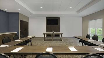 Un salón de conferencias con una larga mesa, sillas, una pantalla de proyector y un ventanal con vistas a zonas verdes.