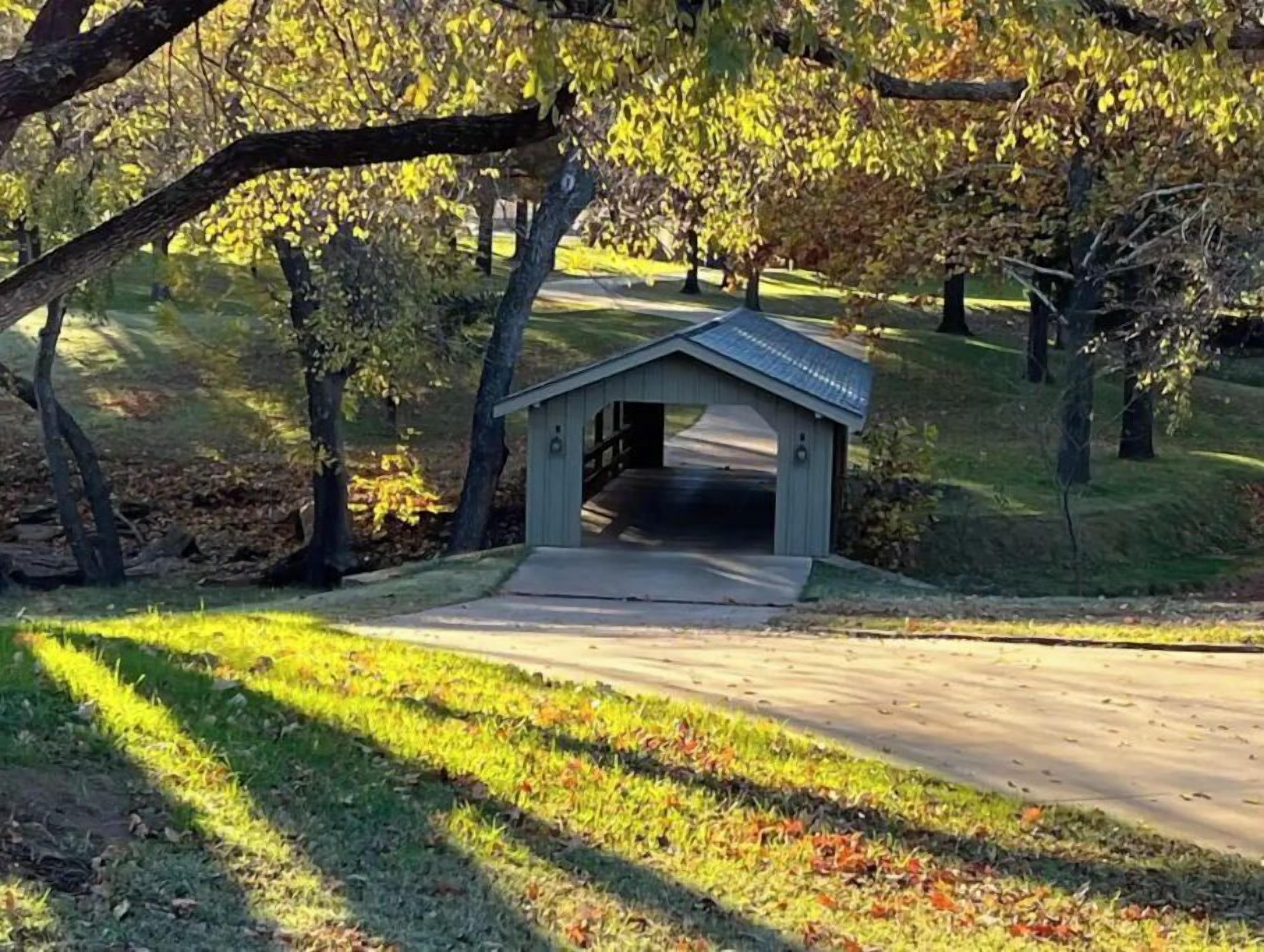 A small covered structure with a roof, likely a shelter or pavilion, situated on a grassy area with a paved path leading to it.