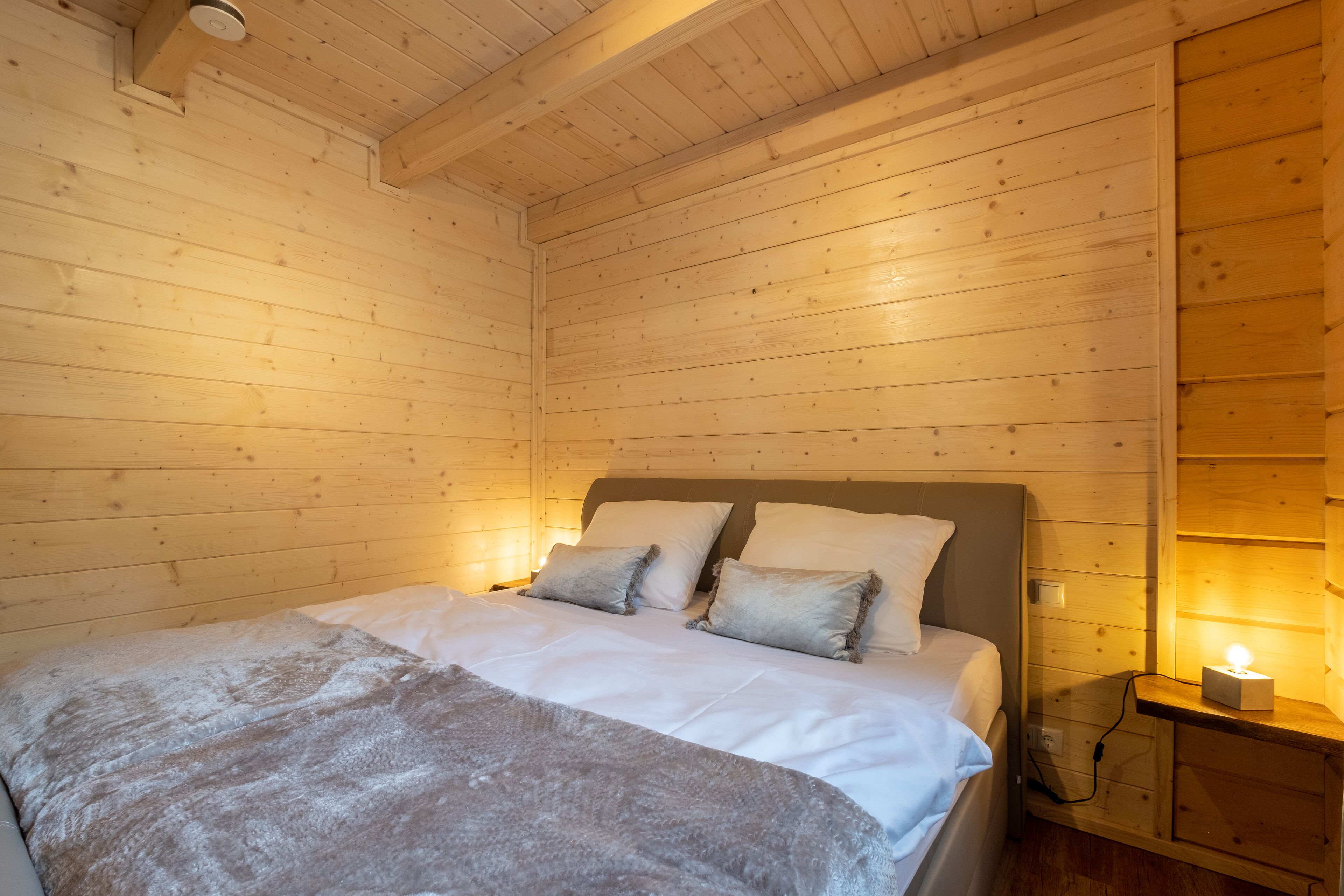Ein Zimmer in einer Holzhütte mit einem Bett, Kissen, einem Nachttisch mit Lampe und einem an der Wand befestigten Regal.