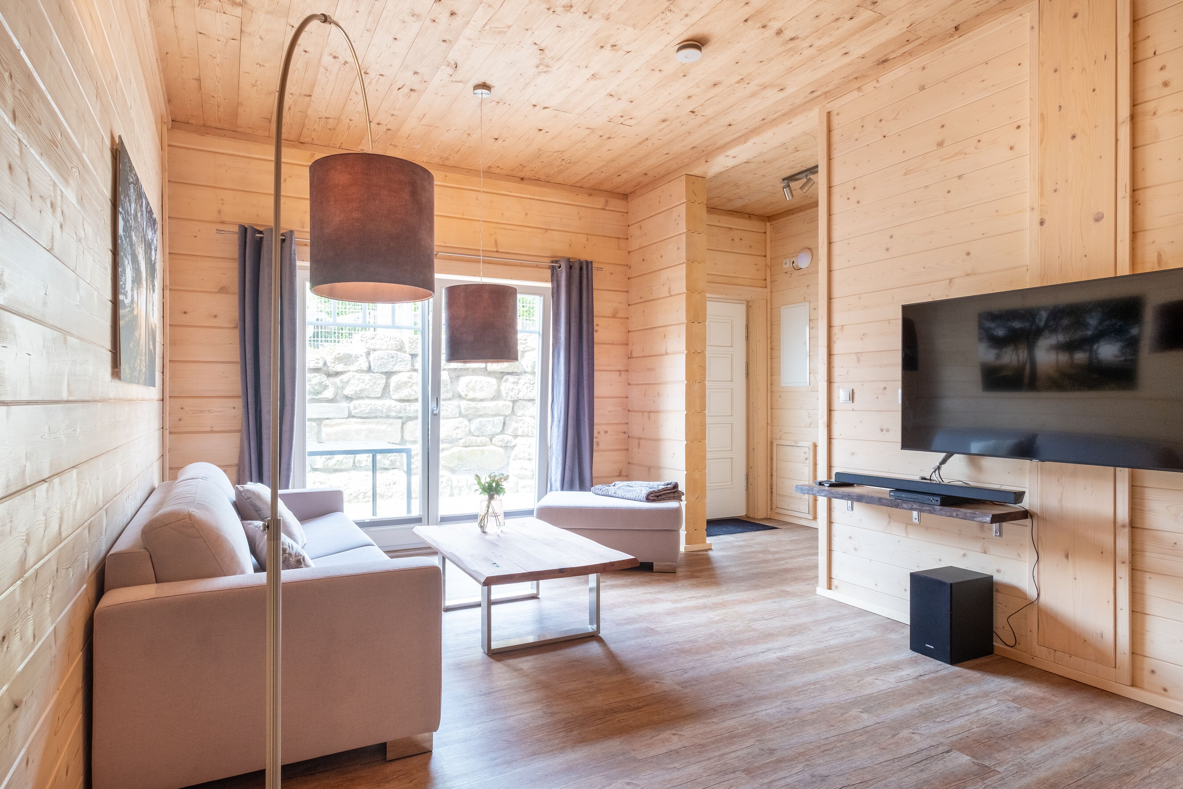 A modern log cabin interior with a sofa, a coffee table, a TV mounted on the wall, and a stone wall visible through the window.