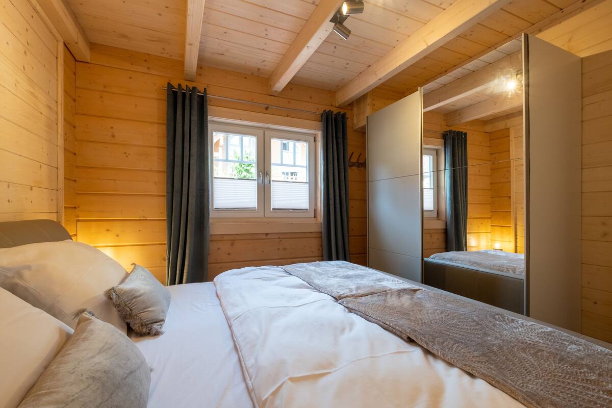 Ein Zimmer mit Holzwänden, einem Bett, einem Fenster mit Vorhängen und einem Spiegel.