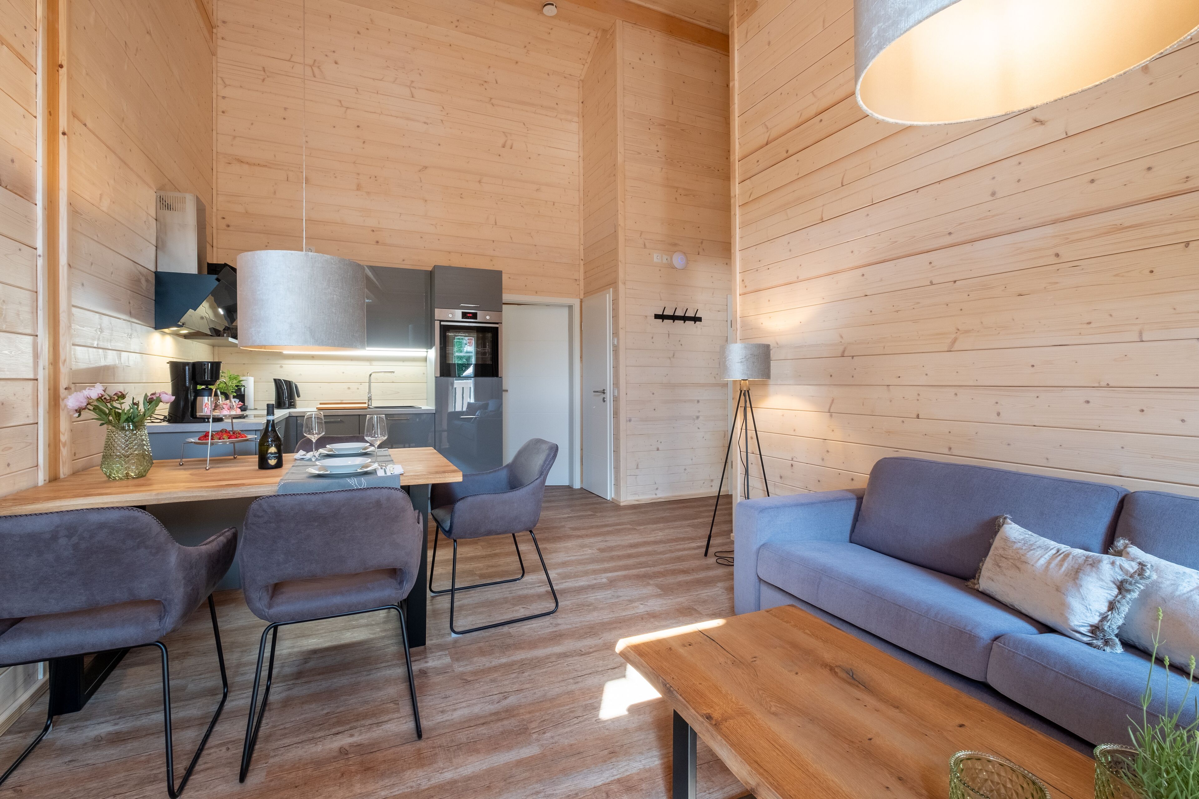 Eine moderne Küche mit Holzwänden, ein Essbereich mit Tisch und Stühlen sowie ein Wohnbereich mit Sofa und Lampe.