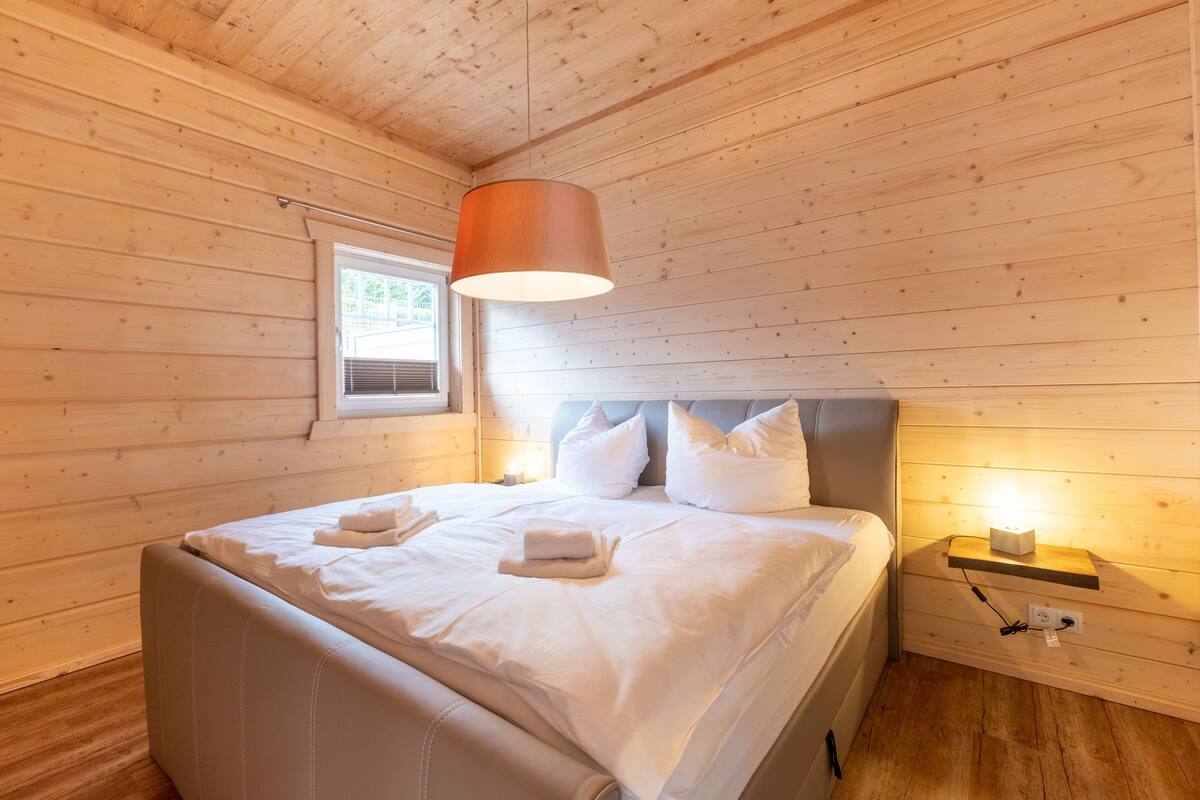 Ein Zimmer mit Holzwänden, einem Bett, Kissen, einem Nachttisch und einem Fenster.