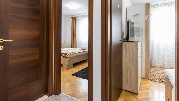 A hotel room with a wooden door, a bed with a patterned bedspread, a desk with a computer, and a television on a stand. - Romeo & Julia (Split)