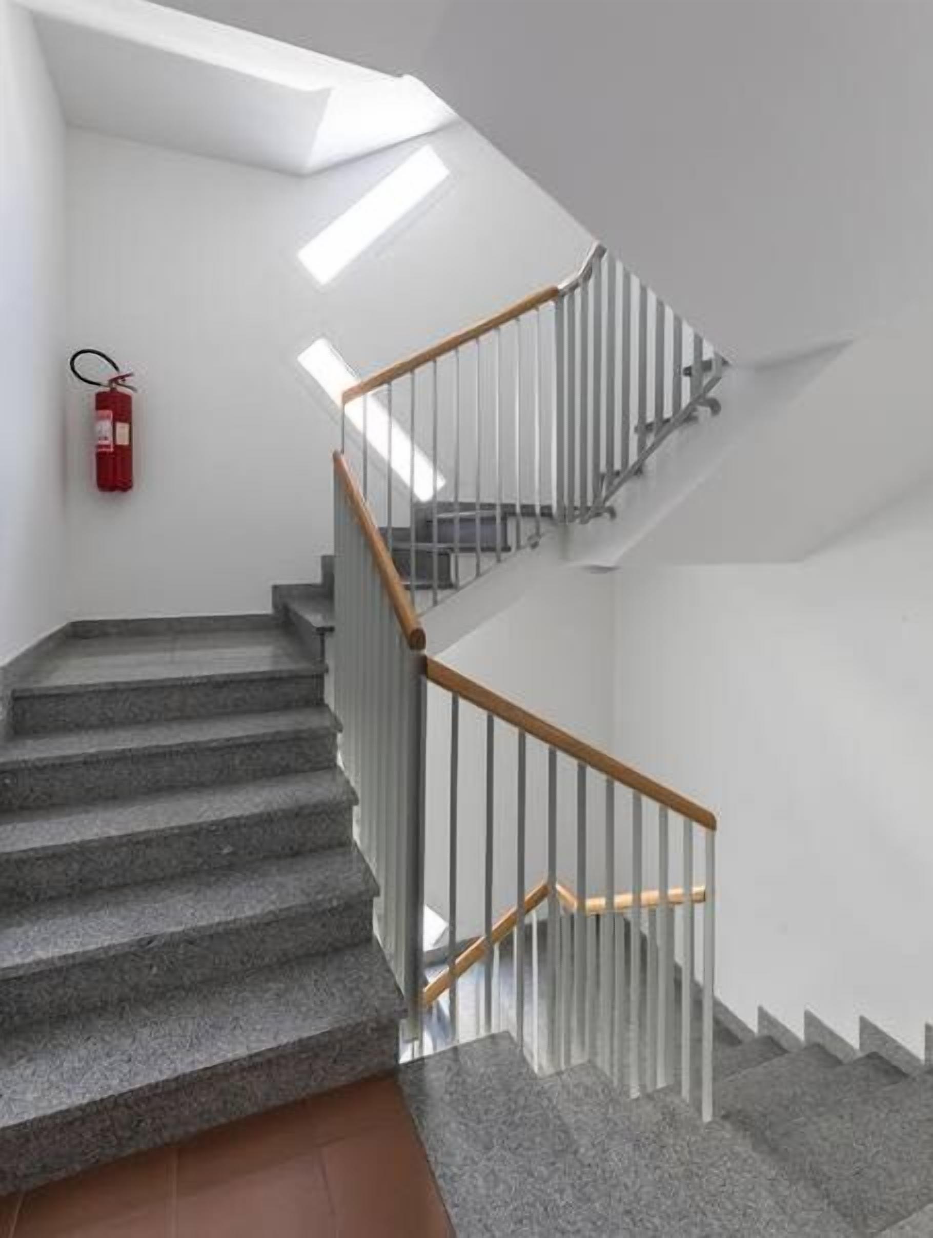 A staircase with stone steps and wooden handrails, leading to a landing with a fire extinguisher on the wall.