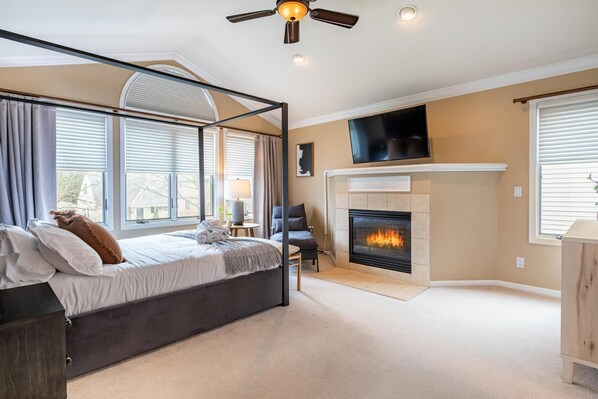 A bedroom with a four-poster bed, a fireplace, a television mounted above it, and large windows with blinds.
