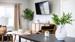 Una sala moderna con una mesa oscura, un jarrón blanco con plantas verdes, una vela pequeña y un televisor en la pared.