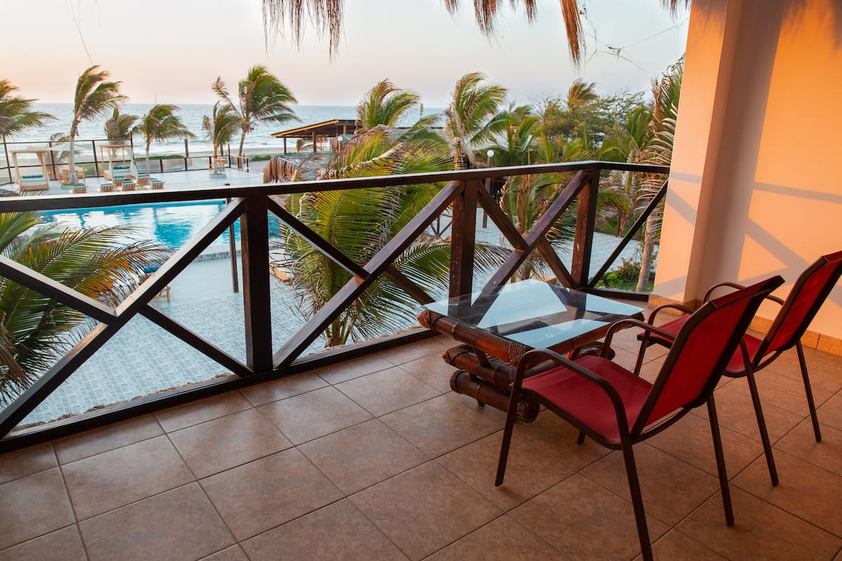 A balcony with a glass railing overlooking a pool and the ocean, furnished with red chairs and a table.