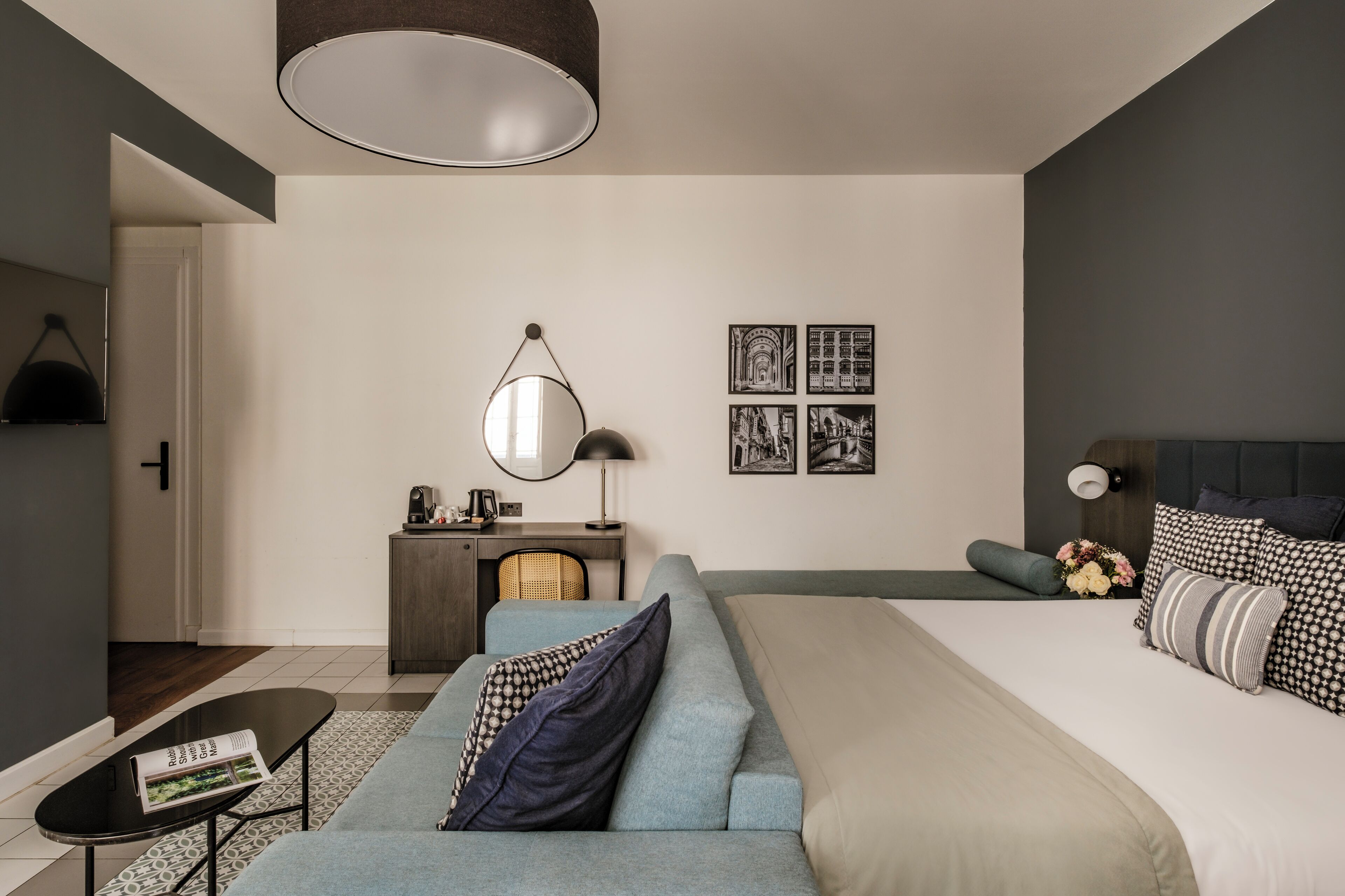 A modern hotel room with a large bed, a sofa, a desk with a lamp, and a wall-mounted mirror.