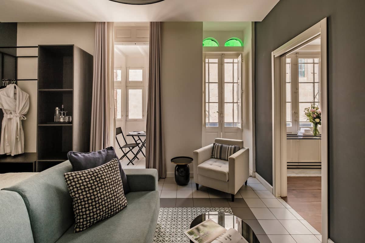 A modern hotel room with a sofa, armchair, and a small side table. There is a bookshelf and a window with curtains.