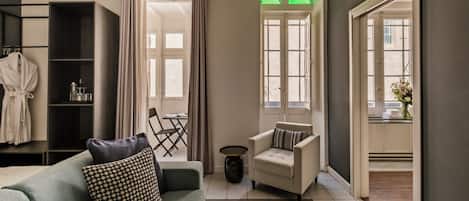A modern hotel room with a sofa, armchair, and a small side table. There is a bookshelf and a window with curtains.