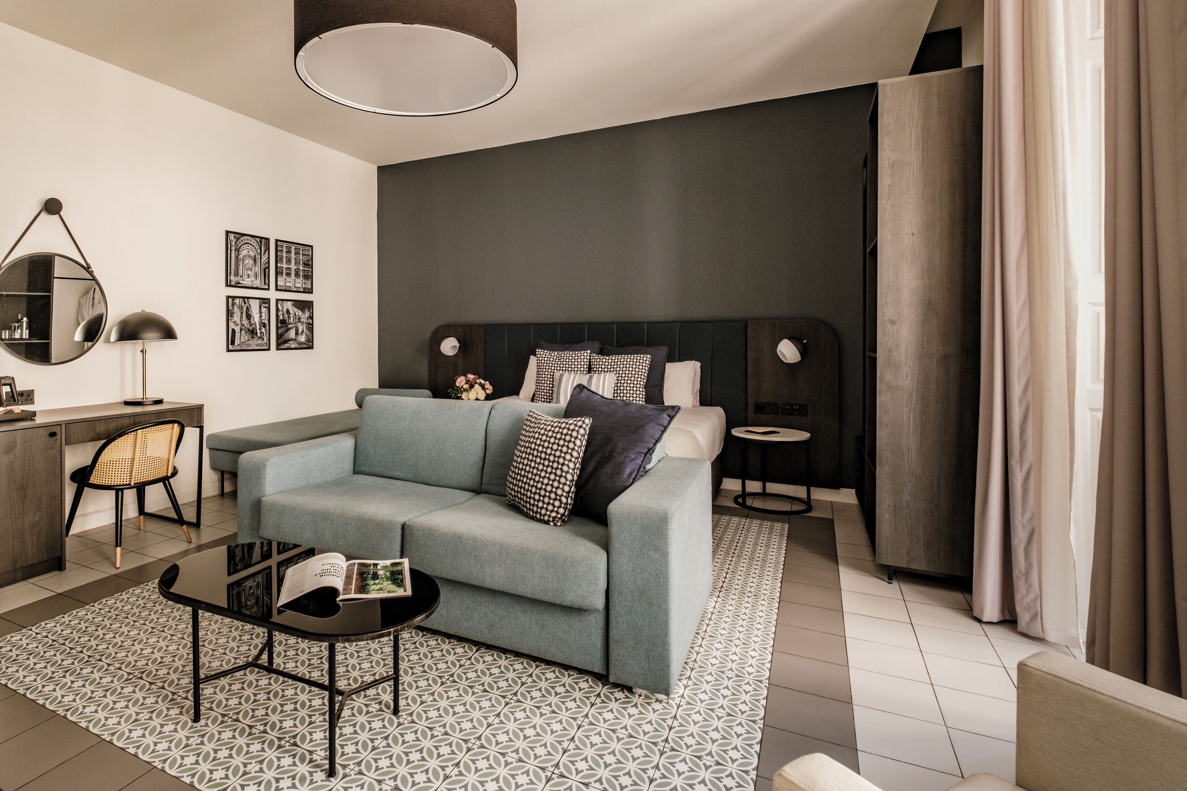 A modern living room with a grey sofa, a coffee table, and a side table. There is a bookshelf, a lamp, and a mirror on the wall.