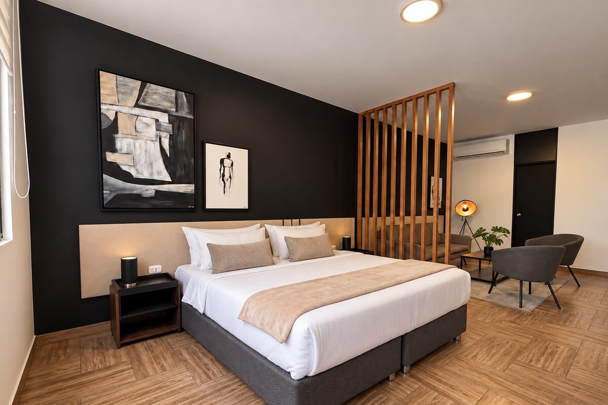 A modern hotel room with a large bed, a bedside table, a desk with a chair, and a large window.