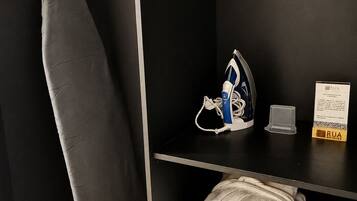 Ein Hotelzimmer mit einem hölzernen Kleiderbügel, einem Bügelbrett, einem Bügeleisen, einem RUA Hotelschild und ordentlich gefalteten Handtüchern auf einem Regal.