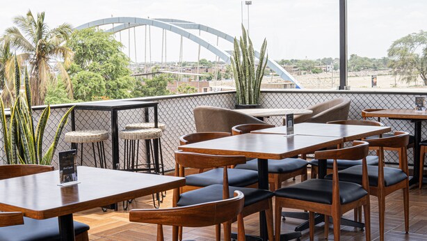 An outdoor dining area with wooden tables and chairs, potted plants, and a view of a bridge and cityscape.