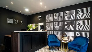 A modern hotel reception area with a dark reception desk, two blue armchairs, and patterned wall decor.