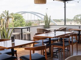 An outdoor dining area with tables and chairs, a view of a bridge, and some greenery.