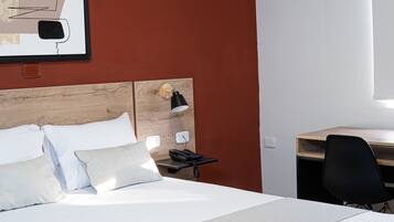 Habitación de hotel con cabecera de madera, cama con sábanas blancas, escritorio con silla y lámpara de pared.