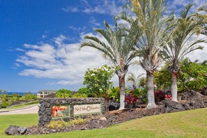 A sign with the text "Na Hale Keauhou" surrounded by tropical landscaping and a view of a building in the background.
