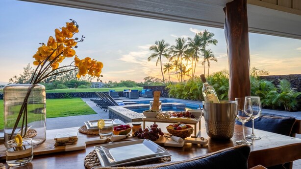 A dining table with a view of a pool and palm trees, a bottle of wine, and various snacks. - Waikoloa Luxe Golf Estate w/ Hilton Pool passes, Gameroom (Puako)