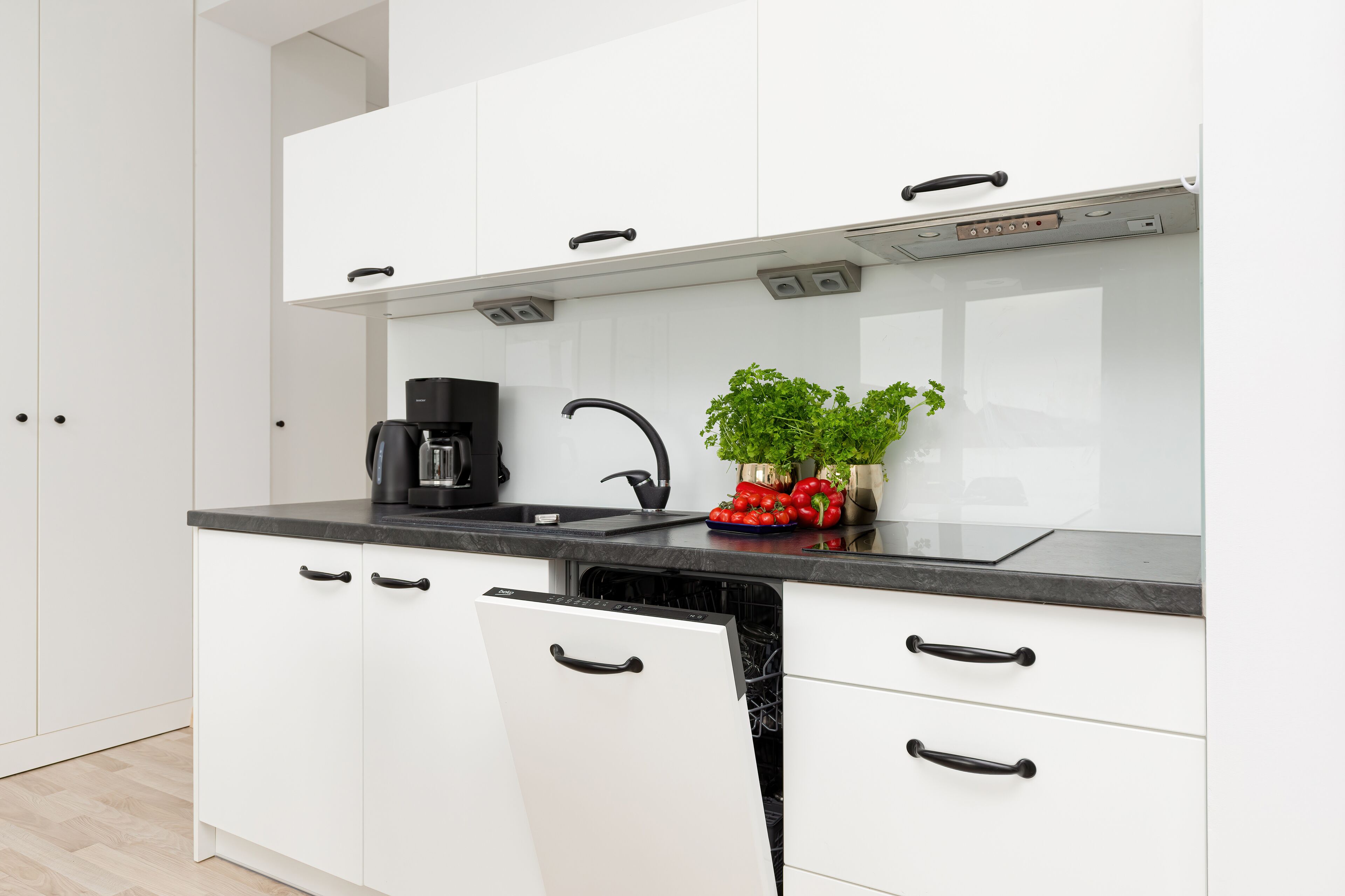 Una cocina moderna con armarios blancos, herrajes negros, una cafetera negra, un lavabo y una planta con pimientos rojos.