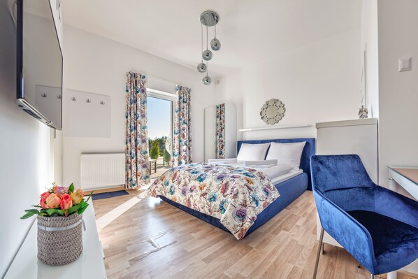 A modern bedroom with a large bed, a blue chair, a small table with a vase of flowers, and a view of the outdoors. - Seaside Apartamenty-Grzybowo  (Kolobrzeg)