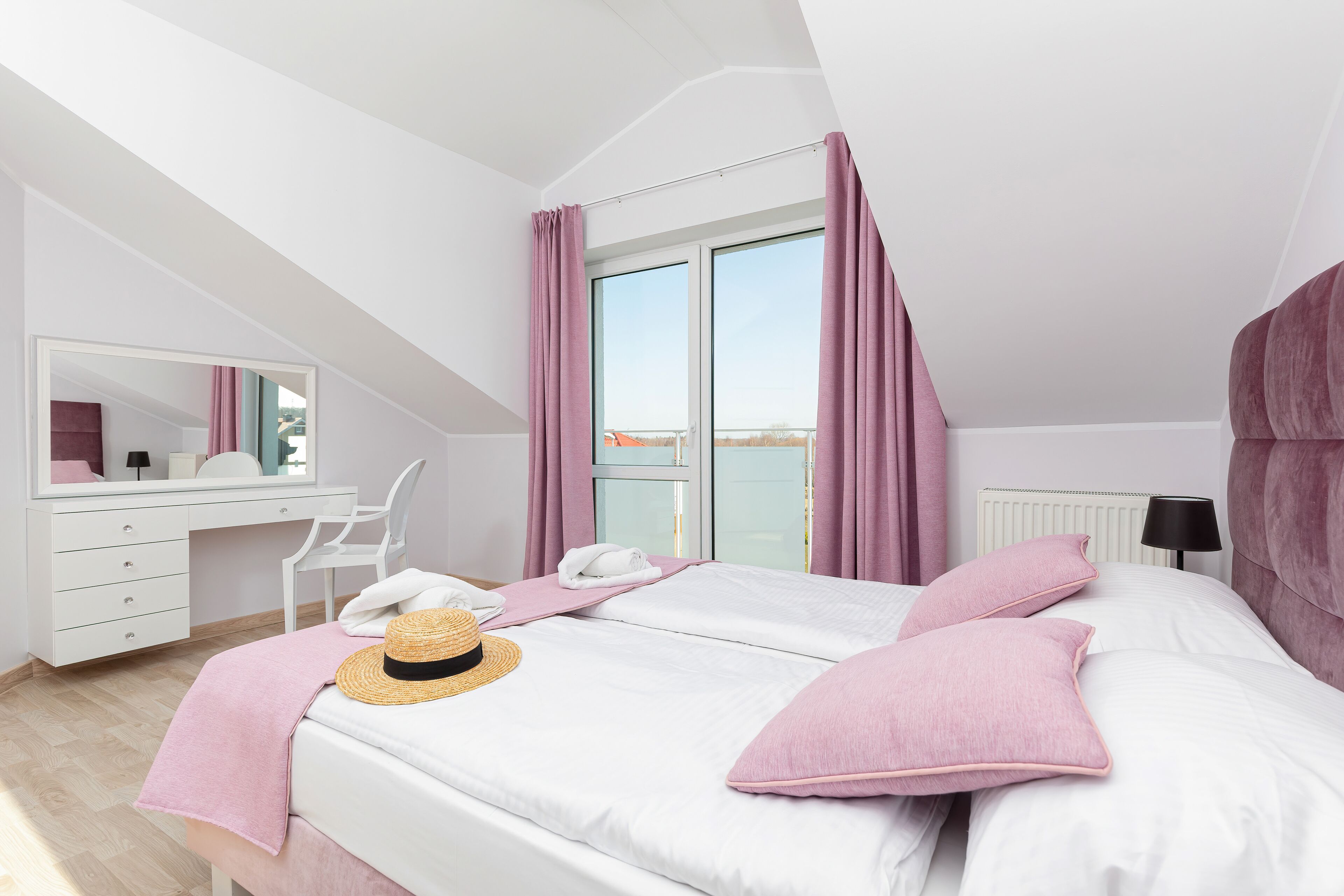 A bedroom with a white bed, pink pillows, a straw hat, and a white desk with a mirror.