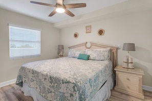Une chambre à coucher avec un lit, un ventilateur de plafond, une table de chevet avec une lampe et une fenêtre avec des stores.