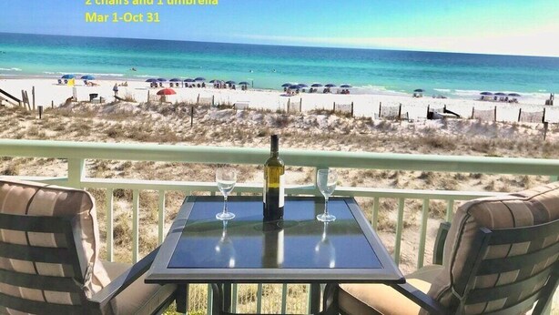 A beach view with a table and chairs, a bottle of wine, and umbrellas. Text indicates beach setup service, 2 chairs, 1 umbrella, available from Mar 1 to Oct 31. - Lovely beach-front condo with free beach setup. (Fort Walton Beach)