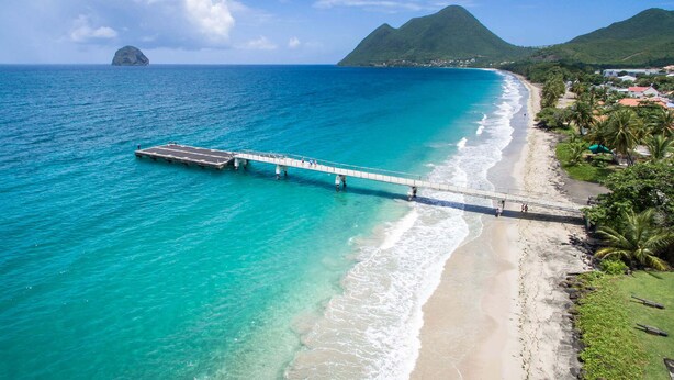 Een kustlandschap met een pier die zich uitstrekt over turquoise water, een zandstrand en weelderige begroeiing.
