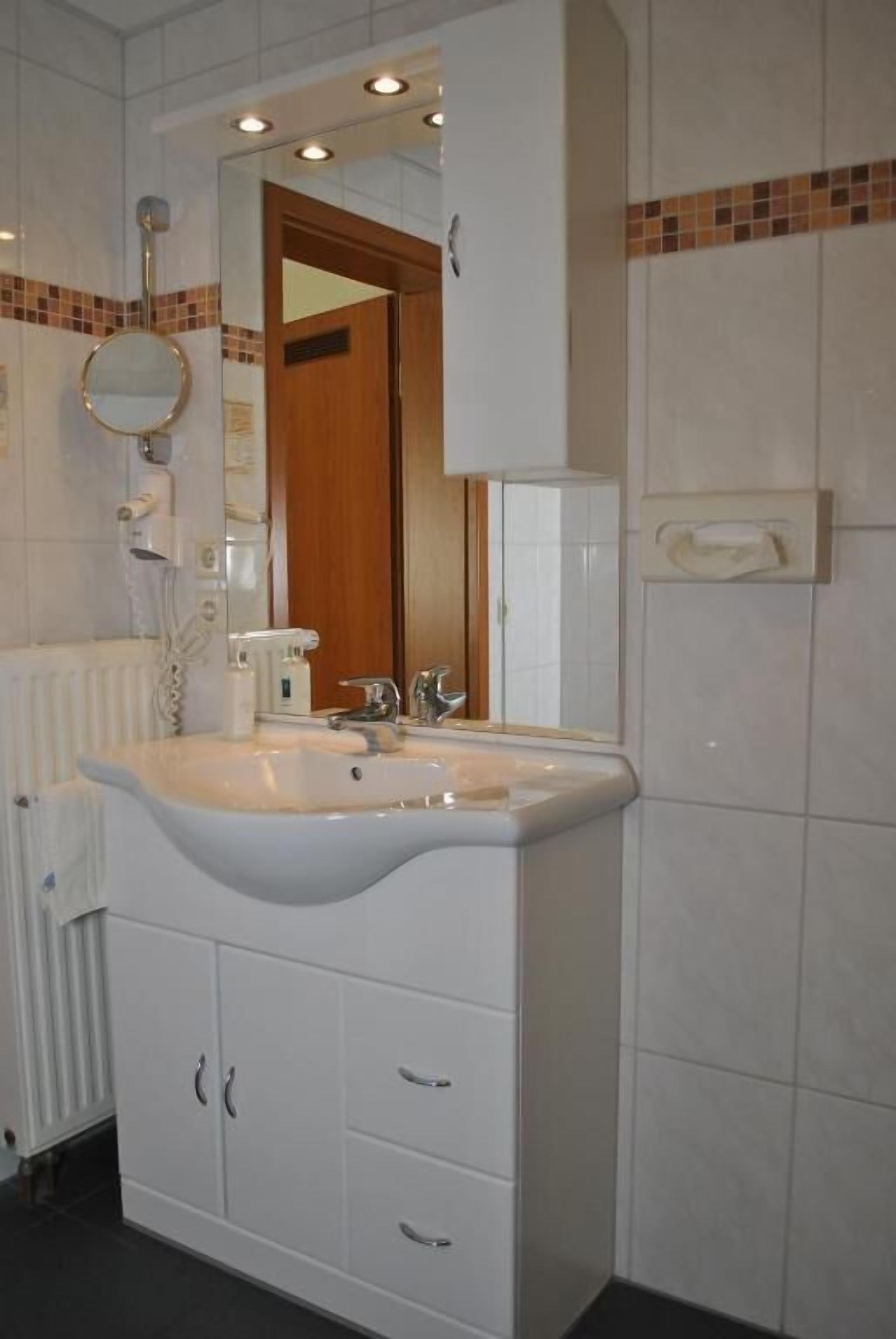 Ein Badezimmer mit weißem Waschbecken, Spiegel und Schrank, einem Handtuchspender und einem Heizkörper.