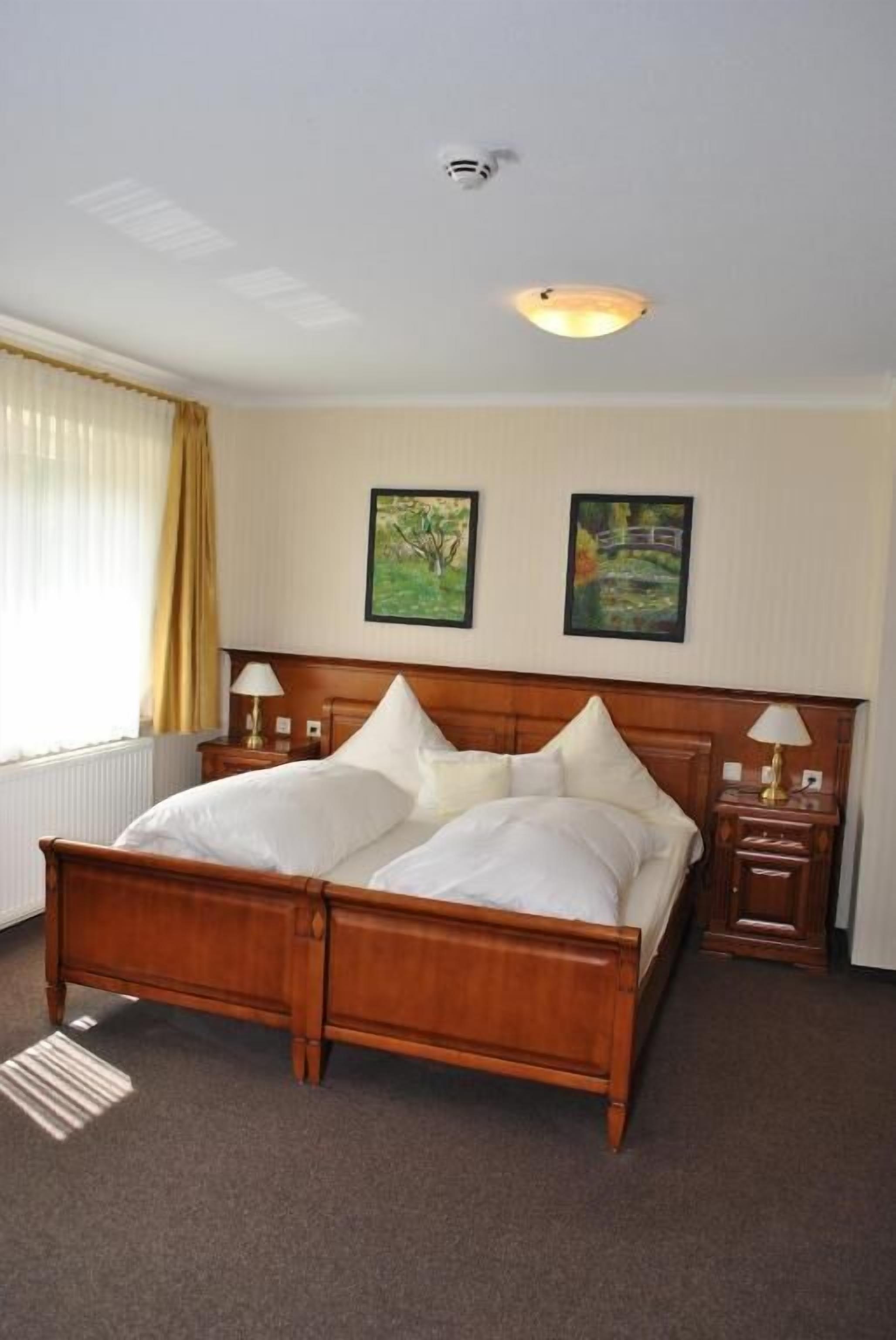 A hotel room with a wooden bed, two bedside tables with lamps, a radiator, and two framed pictures on the wall.