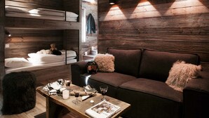 A cozy room with a wooden wall, a sofa with fluffy pillows, a coffee table with drinks and a magazine, and a bunk bed with a teddy bear.