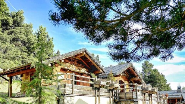 A rustic lodge with stone walls, wooden balconies, and a grassy yard with lounge chairs.