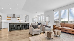 A spacious living area with a kitchen, a brown leather sofa, and a large window with blinds.