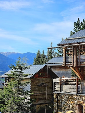 Een rustieke houten chalet met een stenen fundering, een houten balkon en uitzicht op bergen op de achtergrond.