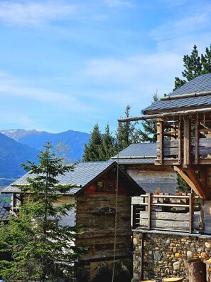 Une cabane rustique en bois, avec des fondations en pierre, un balcon en bois et une vue sur les montagnes en arrière-plan.