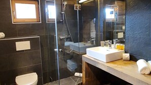 Een moderne badkamer met een glazen douchecabine, een wit toilet, een wastafel met een wasbak en spiegel, en een houten plank met handdoeken.