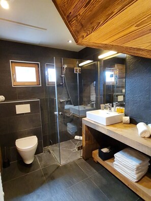 Une salle de bain moderne avec une douche à parois de verre, des toilettes blanches, un meuble-lavabo avec un miroir et un plan de toilette, ainsi qu’une étagère en bois où sont posées des serviettes.