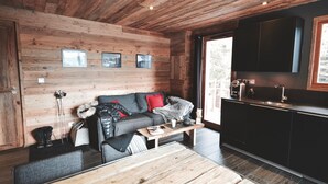 Een gezellig chaletinterieur met houten plafond, een bank met kussens, een bijzettafeltje en een keuken met kastjes en een spoelbak.