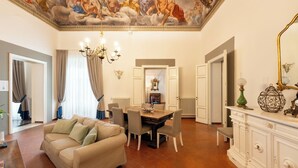 Un'ampia zona giorno con un divano beige, un tavolo da pranzo con sedie, un lampadario e un affresco al soffitto.