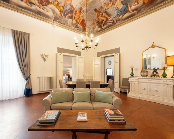 Un salon spacieux avec un canapé beige, une table basse sur laquelle sont posés des livres, un lustre et une fresque au plafond.